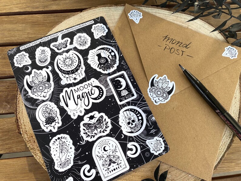 Puede incluir: Una hoja de pegatinas en blanco y negro con las palabras "Moon Magic" y varias ilustraciones con tem&aacute;tica lunar, de cristales y celestial. La hoja est&aacute; sobre una superficie de madera.