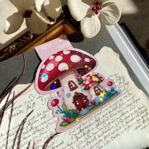 Puede incluir: Un marcapáginas holográfico rosa con una casa de seta roja y blanca y una pequeña casa de seta blanca a su lado. Las casas de seta tienen un interior azul y blanco y una puerta marrón. El marcapáginas está decorado con purpurina iridiscente.