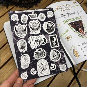Puede incluir: Una hoja de pegatinas con ilustraciones en blanco y negro de fases lunares, cristales, mariposas y otros elementos m&aacute;gicos. La hoja de pegatinas est&aacute; sobre un fondo negro con estrellas blancas. El texto "Moon Magic" est&aacute; impreso en blanco sobre una pancarta negra. La hoja de pegatinas se titula "Sticker Sheet Moon Magic".