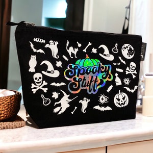 Könnte beinhalten: Schwarze Kosmetiktasche mit Reißverschluss und weißen Halloween-Motiven wie Geistern, Fledermäusen und einer Hexe. Die Tasche trägt den Schriftzug "Spooky Stuff" in einem holografischen Design.
