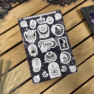 Puede incluir: Una hoja de pegatinas con un dise&ntilde;o en blanco y negro que presenta varias ilustraciones con tem&aacute;tica lunar y celestial. La hoja incluye una calavera, una luna creciente, un gato, una mariposa y un cristal. El texto "Moon Magic" aparece en la hoja.
