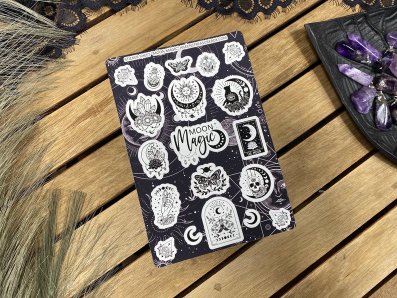 Puede incluir: Una hoja de pegatinas con un dise&ntilde;o en blanco y negro que presenta varias ilustraciones con tem&aacute;tica lunar y celestial. La hoja incluye una calavera, una luna creciente, un gato, una mariposa y un cristal. El texto "Moon Magic" aparece en la hoja.