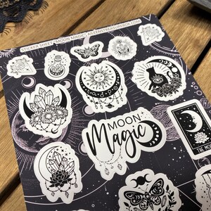 Puede incluir: Una hoja de pegatinas con un tema de magia lunar en blanco y negro. La hoja presenta varios dise&ntilde;os, incluyendo una luna creciente, un gato, una calavera, una mariposa, cristales y un sol. El texto "MOON MAGIC" est&aacute; impreso en la hoja.