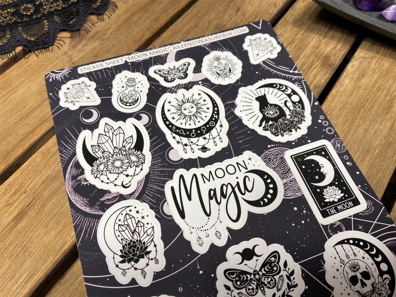 Puede incluir: Una hoja de pegatinas con un tema de magia lunar en blanco y negro. La hoja presenta varios dise&ntilde;os, incluyendo una luna creciente, un gato, una calavera, una mariposa, cristales y un sol. El texto "MOON MAGIC" est&aacute; impreso en la hoja.