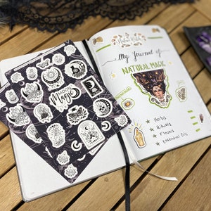 Puede incluir: Una hoja de pegatinas en blanco y negro con las palabras "Moon Magic" en ella. Las pegatinas presentan varias fases lunares, cristales y otros s&iacute;mbolos de brujer&iacute;a. Las pegatinas est&aacute;n sobre un fondo blanco y se est&aacute;n colocando en un diario con las palabras "My Journal of Natural Magic" escritas en la p&aacute;gina.