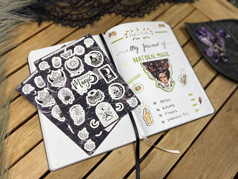 Puede incluir: Una hoja de pegatinas en blanco y negro con las palabras "Moon Magic" en ella. Las pegatinas presentan varias fases lunares, cristales y otros s&iacute;mbolos de brujer&iacute;a. Las pegatinas est&aacute;n sobre un fondo blanco y se est&aacute;n colocando en un diario con las palabras "My Journal of Natural Magic" escritas en la p&aacute;gina.