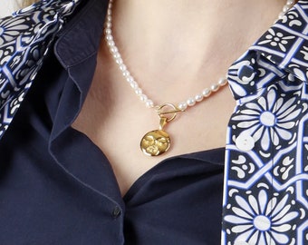 May include: A white pearl necklace with a gold pendant featuring a face design. The necklace is worn by a person wearing a navy blue shirt with a white and blue patterned design.