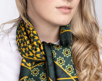 May include: A woman wearing a green and gold patterned scarf with a geometric design. The scarf is tied around her neck and the ends are hanging down. She is wearing a white shirt underneath the scarf. She is also wearing a pair of earrings with a green gemstone.