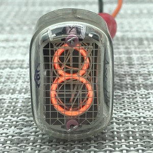 Könnte beinhalten: Nahaufnahme einer Vintage-Nixie-Röhre, die die Zahl 8 in leuchtendem Orange anzeigt. Die Röhre ist in klarem Glas mit einer gitterartigen Struktur im Inneren eingeschlossen. Drähte erstrecken sich von oben, und die Röhre befindet sich auf einer strukturierten grauen Oberfläche.