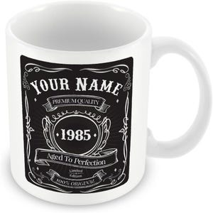 Pu&ograve; includere: Tazza in ceramica bianca con un design in bianco e nero. Il design presenta il testo "YOUR NAME", "PREMIUM QUALITY", "1985", "Aged To Perfection", "Limited Edition" e "100% ORIGINAL".