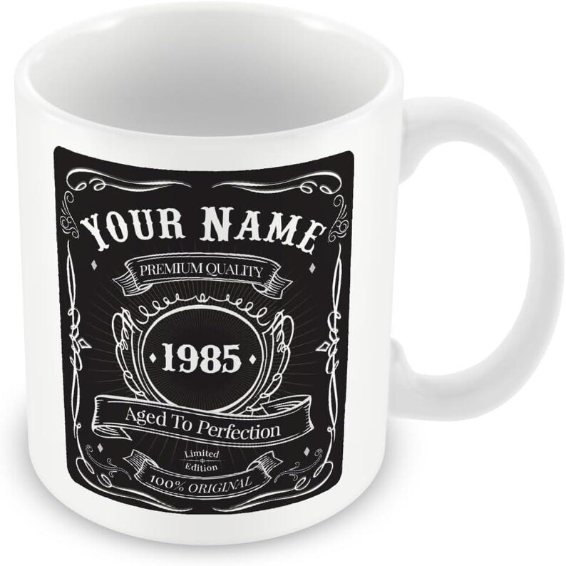 Pu&ograve; includere: Tazza in ceramica bianca con un design in bianco e nero. Il design presenta il testo "YOUR NAME", "PREMIUM QUALITY", "1985", "Aged To Perfection", "Limited Edition" e "100% ORIGINAL".