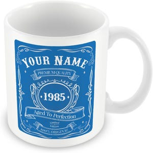 Pu&ograve; includere: Tazza in ceramica bianca con un design blu. Il design presenta un'etichetta vintage con il testo "YOUR NAME", "PREMIUM QUALITY", "1985", "Aged To Perfection", "Limited Edition" e "100% ORIGINAL".