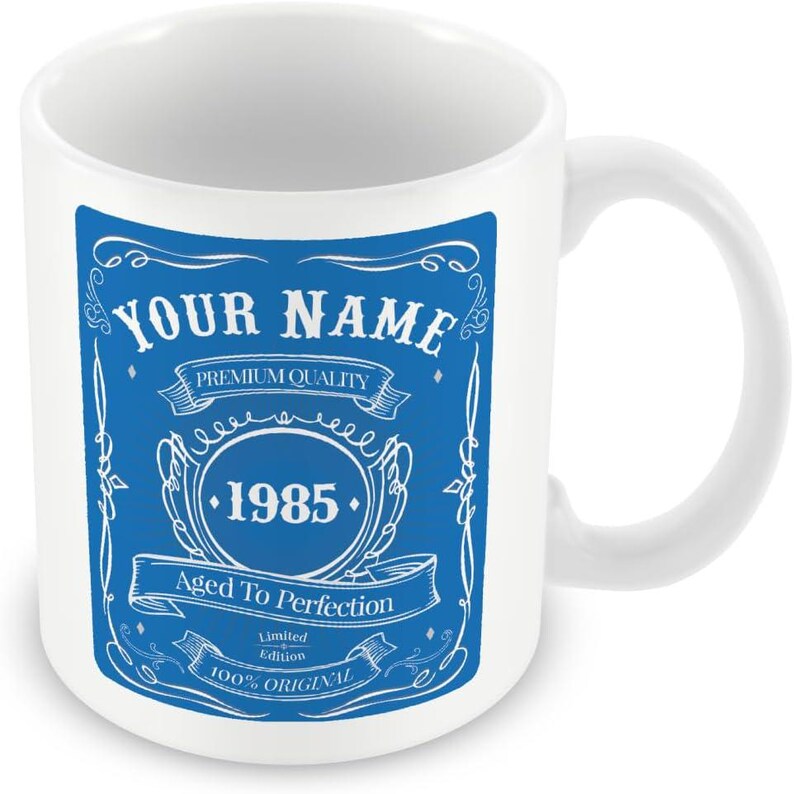 Pu&ograve; includere: Tazza in ceramica bianca con un design blu. Il design presenta un'etichetta vintage con il testo "YOUR NAME", "PREMIUM QUALITY", "1985", "Aged To Perfection", "Limited Edition" e "100% ORIGINAL".