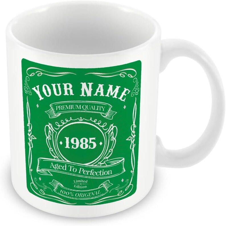 Pu&ograve; includere: Tazza in ceramica bianca con un design verde. Il design presenta il testo "YOUR NAME", "PREMIUM QUALITY", "1985", "Aged To Perfection", "Limited Edition" e "100% ORIGINAL".