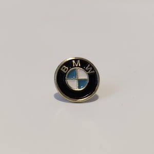 Pode incluir: Um alfinete dourado com um logotipo BMW preto e branco. O logotipo é um círculo com um design de hélice branco e azul dentro. As letras "BMW" são escritas em torno do círculo em preto.