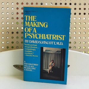 May include: A blue book with the title "The Making of a Psychiatrist" by David S. Viscott, M.D. The book cover features a photo of a woman standing in a room with a window.
