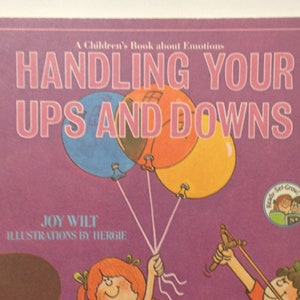 May include: A children's book titled "Handling Your Ups and Downs" with illustrations of two children, one holding balloons and the other holding a slingshot. The book cover is purple with a yellow, orange, and blue balloon design.