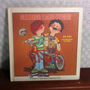 May include: A brown book with the title "Needing Each Other" on the cover. The cover features an illustration of two children riding a bicycle together. The book is a children's book about relational needs.