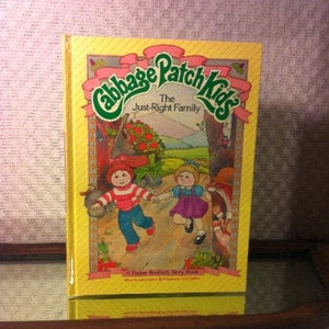 May include: A yellow book with a green and pink Cabbage Patch Kids logo. The title is "The Just-Right Family" and the book is a "Pocket Edition Story Book". The cover features illustrations of two Cabbage Patch Kids dolls, one with red hair and a red and white striped shirt, and the other with blonde hair and a purple dress.