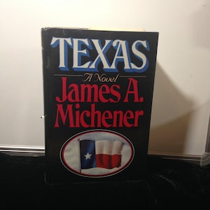 May include: A black hardcover book with the title "TEXAS" in white and blue lettering. The book is by James A. Michener and features a red, white, and blue Texas flag on the cover.