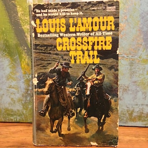 May include: A vintage paperback book titled "Crossfire Trail" by Louis L'Amour. The cover features a Western scene with cowboys on horseback. The book's title and author's name are in bold yellow lettering against a backdrop of a Western landscape.