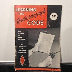 May include: A vintage book titled "Learning the Radiotelegraph Code" with a red and black cover. The book is published by the American Radio Relay League and costs 50 cents. The cover features a person using a radiotelegraph key.