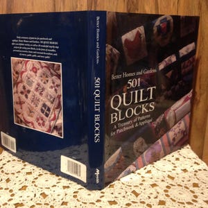 May include: A hardcover book titled "501 Quilt Blocks" by Better Homes and Gardens. The book features a navy blue spine with white text and a colorful cover showcasing quilt block designs. The book is displayed on a white lace surface.