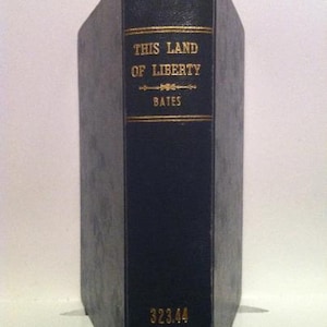 May include: A vintage book with a dark blue cover and gold lettering. The title "THIS LAND OF LIBERTY" and the author's name "BATES" are displayed. The book's spine also features the number "323.44".