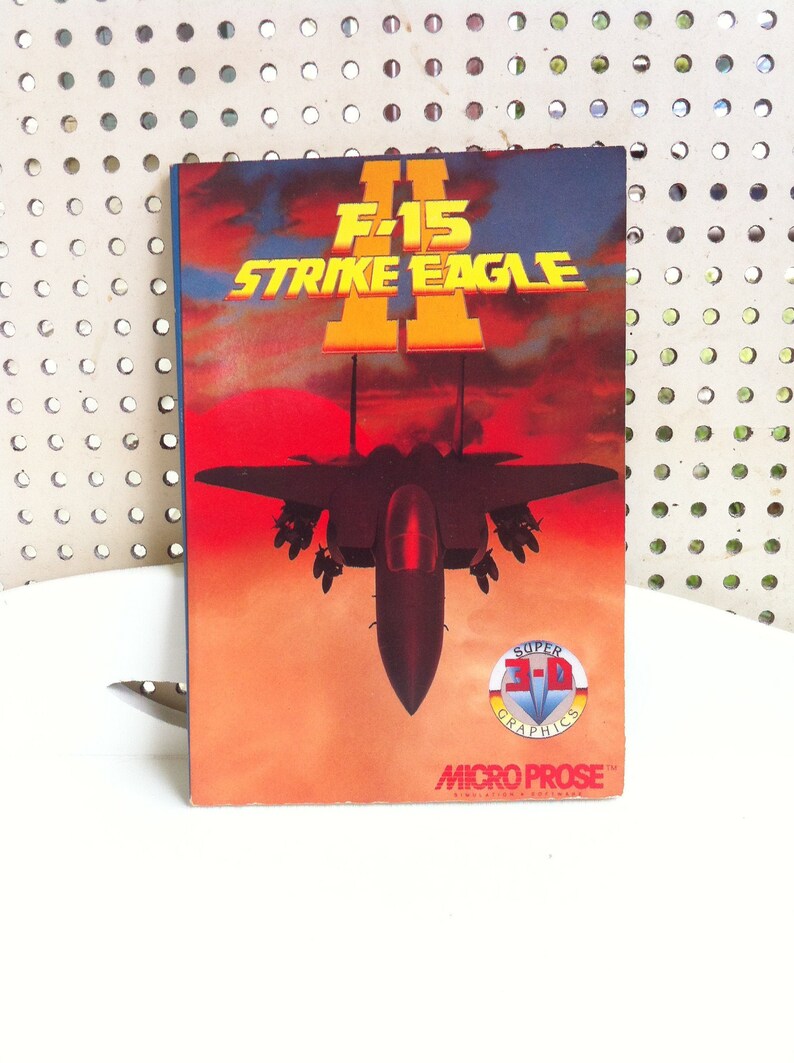 May include: A vintage video game box cover for F-15 Strike Eagle II, featuring a black fighter jet against a red and orange sunset sky. The box features the text "Super 3-D Graphics" and the MicroProse logo.