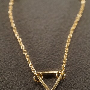 May include: A gold-colored necklace featuring a delicate chain and a geometric triangle pendant. The pendant is a simple, open-frame design. The necklace is displayed against a textured, dark gray background.