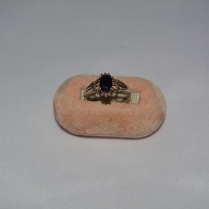 May include: A gold ring with a dark blue oval gemstone surrounded by smaller clear gemstones. The ring is displayed on a pink velvet cushion.