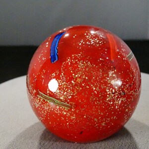 May include: A round, red glass paperweight with gold flecks and colorful accents. The decorative sphere features a blue stripe and a green and gold swirl design. This art glass piece is a unique home decor item.