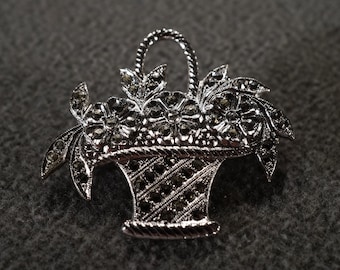 Puede incluir: Un broche de plata en forma de cesta con un diseño floral. La cesta está llena de flores y hojas negras y plateadas.