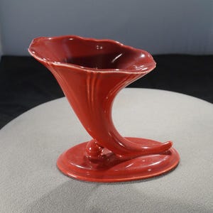 May include: A red ceramic vase shaped like a horn. The vase has a wide, flared opening and a narrow base.