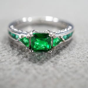 May include: A silver ring with a large, square, emerald-green gemstone at the center, flanked by two smaller triangular green stones. The band is adorned with smaller green stones and clear accents.