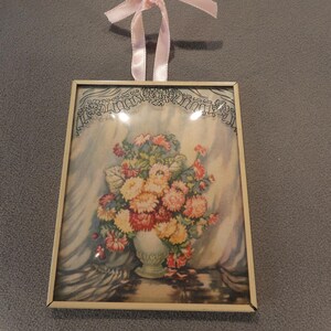 May include: A rectangular wall hanging featuring a floral print in a thin gold-tone metal frame. The artwork depicts a bouquet of yellow, orange, and red flowers in a vase against a draped background. A light pink ribbon is attached for hanging.