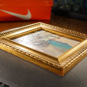 May include: A small framed painting with a gold frame. The painting depicts a landscape with a blue sky, white clouds, and a green field.