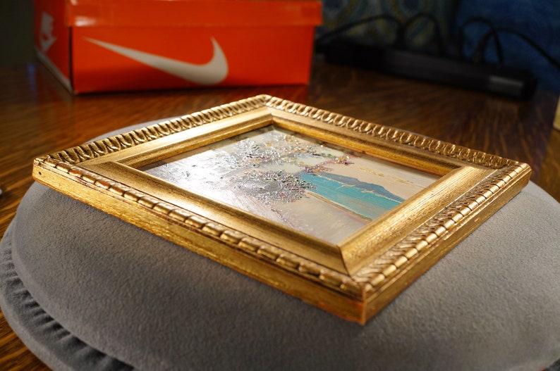 May include: A small framed painting with a gold frame. The painting depicts a landscape with a blue sky, white clouds, and a green field.