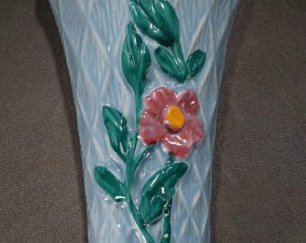 May include: A light blue ceramic wall pocket vase with a diamond pattern. A raised floral design features a red flower with a yellow center and green leaves. The vase has a woven-like trim around the top.