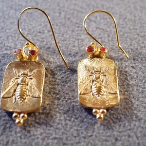 May include: Gold earrings with a bee design and red gemstone accents.
