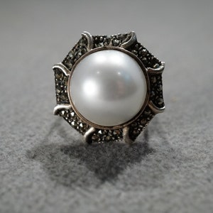May include: A silver-toned ring featuring a large, lustrous white pearl at its center. The pearl is surrounded by a decorative setting with small, dark accents, creating a striking contrast. The ring has an octagonal shape.