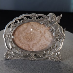 May include: Silver-toned oval picture frame with an ornate design and a cherub figure holding a trumpet. The frame has a light brown background.