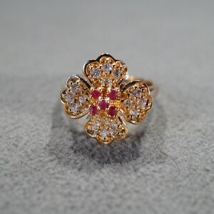 May include: A gold ring with a four-leaf clover design. The clover is set with small clear stones and five red stones in the center.