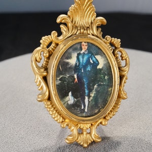 May include: A gold-colored ornate frame with a portrait of a young person wearing a blue suit and white shirt. The portrait is set against a dark background with a white dog in the foreground.