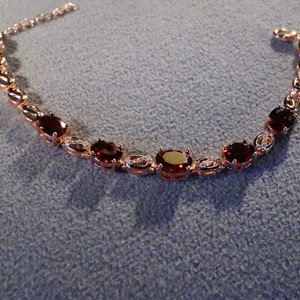 May include: A rose gold bracelet with oval-shaped red gemstones and smaller clear gemstones set in the metal.
