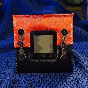 May include: A small, black and orange picture frame with a decorative design. The frame has a square opening for a photo and is decorated with black and gold ribbon.
