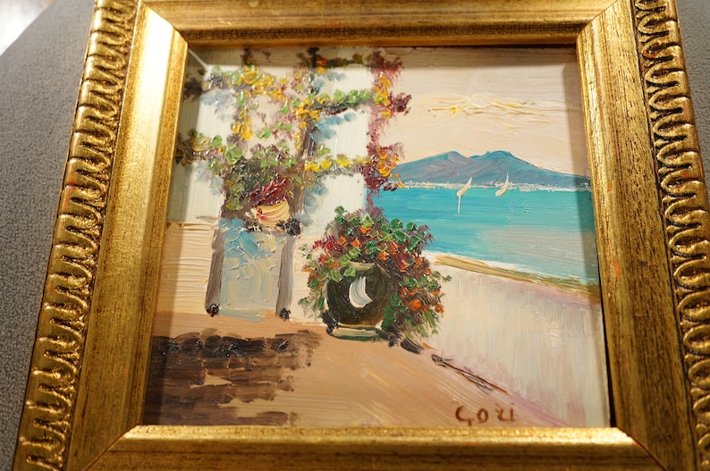 May include: A framed oil painting depicting a seaside scene with a view of a mountain range in the distance. The painting features two potted plants, a white building, and a blue sea with sailboats. The artist's signature, "Gozi", is visible in the lower right corner.