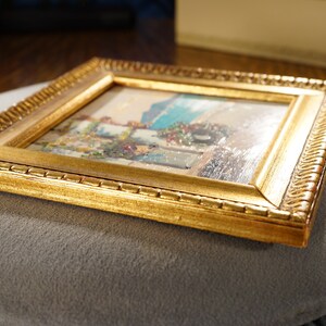 May include: A small framed painting of a coastal scene with a blue sky, white buildings, and a green bush. The frame is gold with a decorative border.