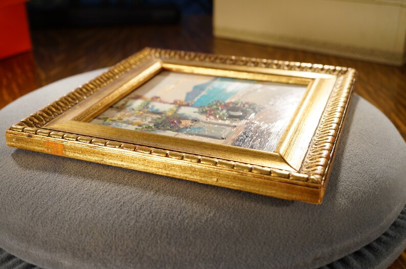 May include: A small framed painting of a coastal scene with a blue sky, white buildings, and a green bush. The frame is gold with a decorative border.
