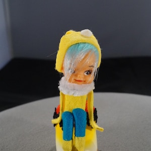May include: A vintage doll with a yellow hat and outfit, blue pants, and white collar. The doll has blue hair and is sitting on a white and yellow base. The doll's face has a smiling expression.